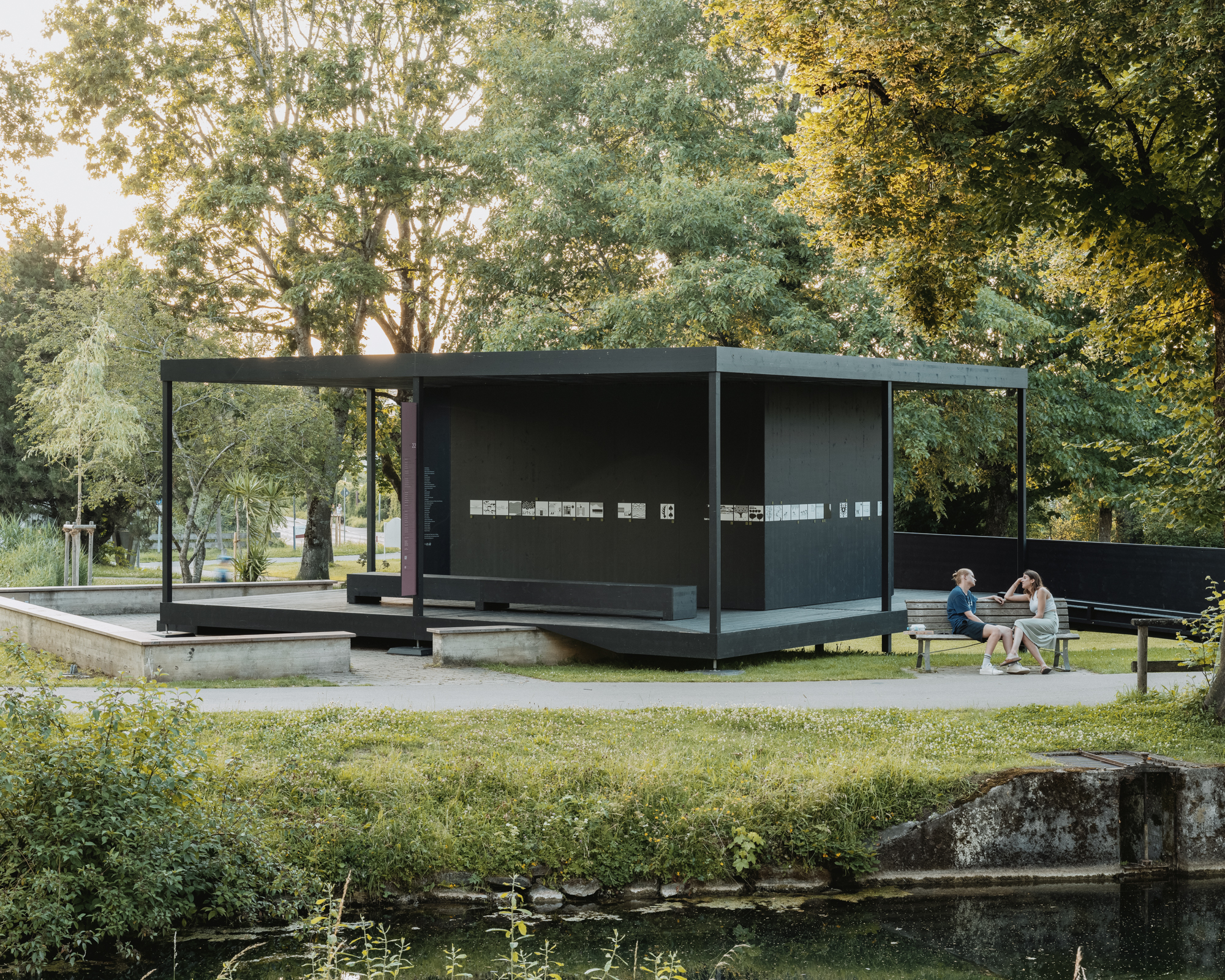 Der temporäre Pavillon besteht aus einer schwarz gestrichenen Holzkonstruktion auf filigranen Stützen. Eine umlaufende Pergola fasst den Ausstellungsraum, dessen offenes Boden- und Deckengitter den Wetterwechsel spürbar macht. Ohne Eingang, Tür oder Öffnungszeiten ist der Pavillon jederzeit und für alle zugänglich – für eine begrenzte Zeit.